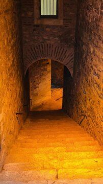 Vertical view of ancient stone steps and brick archway leading down to underground cellar inside historic building. Medieval masonry tunnel walkway with warm iron lanterns light illumination.