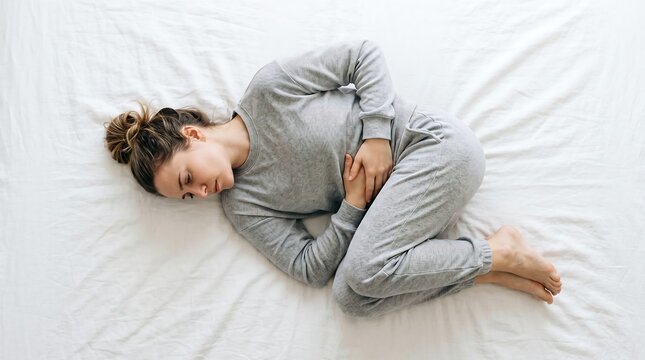 Young woman lying in bed curled up in fetal position suffering from severe stomach pain or menstrual cramps.