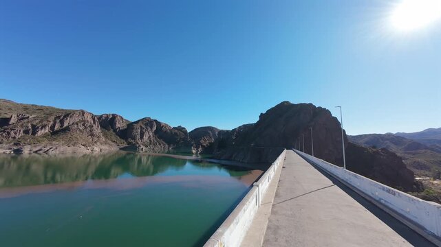 Adult and child walk in Valle Grande Dam in Atuel Canyon, San Rafael, Mendoza, Argentina, overlooking Embalse Valle Grande, hydroelectric infrastructure and surrounding canyon geology, low FPV forward