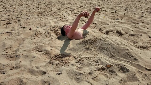 Angle on young boy, half buried in sand as he relaxes and pours sand on himself in the summer sun