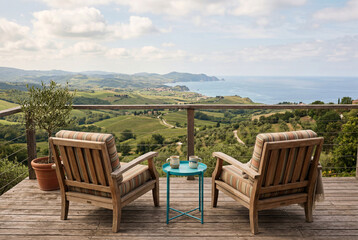 Obraz premium Two empty wooden armchairs on a rustic deck overlooking a green hilly landscape and a blue ocean