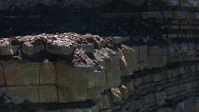 Close-up view of a quarry rock face with exposed geological layers, fractured stone blocks, and loose rubble. The excavated cliff reveals sedimentary strata and active mineral extraction.