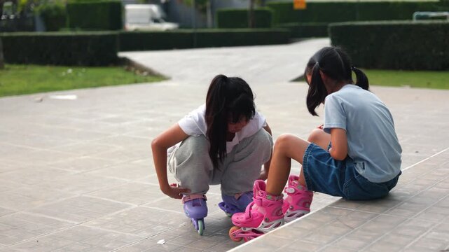 Two happy Asian kids girls learning how to rollerskate together inside green public park courtyard. Cute young classmates sharing friendship outdoor lifestyle clip project.