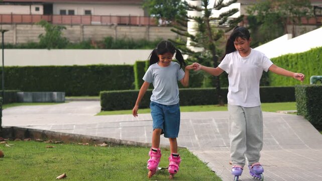 Two active elementary Asian girls teaching rollerskate sport hobby to companion inside public park track. Happy child friends enjoying recreation fitness training spot session.