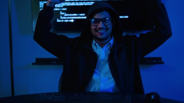 Male hacker in a black hoodie working on a computer in a dark room with blue neon light. He celebrates a successful cyber attack or coding breakthrough. Technology and cyber security concept.