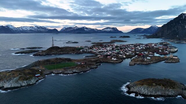 Wide Aerial Drone footage of Henningsvaer Fishing Village and Football pitch with Snowy Mountains, Lofoten, Norway, at sunset, Horizontal lateral shot
