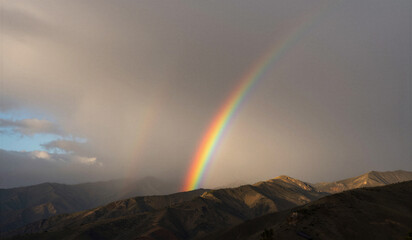 Naklejka premium Rainbow arches gracefully over rugged mountains. Clouds swirl behind, softening the sky's edge. Distant peaks stand bold beneath the natural display. Nature's calm beauty after stormy weather