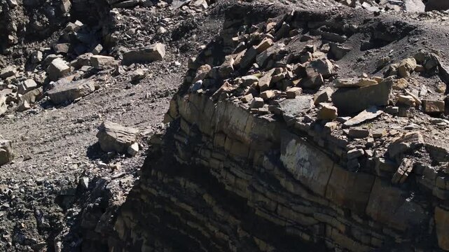 Close-up view of a quarry rock face with exposed geological layers, fractured stone blocks, and loose rubble. The excavated cliff reveals sedimentary strata and active mineral extraction.