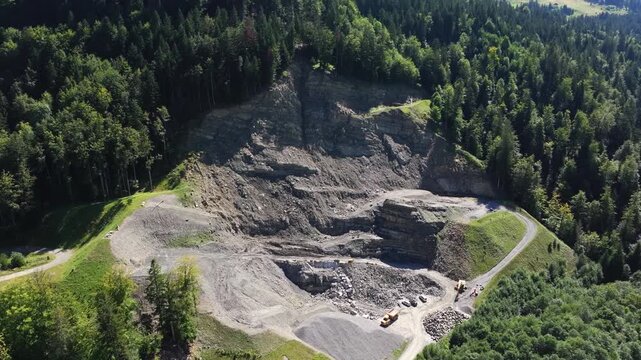Aerial view of an active quarry surrounded by forest. Exposed rock faces, excavation terraces, gravel stockpiles, access roads, and heavy machinery highlight industrial mineral extraction operations.