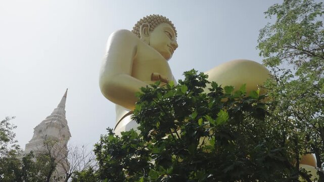 A towering golden Buddha statue rises above lush greenery at a historic temple complex in Bangkok, Thailand. Accompanied by a traditional white stupa, the scene captures the spiritual beauty and archi