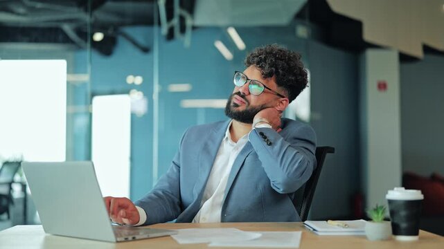 Middle Eastern businessman in a suit rubbing his sore neck in pain. Image depicting muscle tension, office fatigue, and physical discomfort while working at a desk in a modern corporate office.