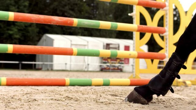 Close up slow motion footage of horse legs jumping over colorful obstacle in equestrian arena dust flying during event.
