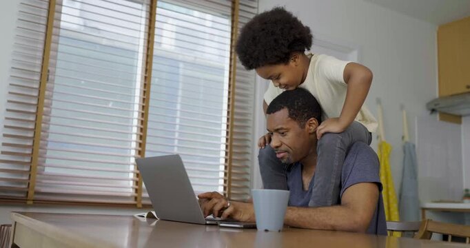 Busy father using a laptop while young child plays on his shoulders, illustrating remote work, parenting challenges, and family life.