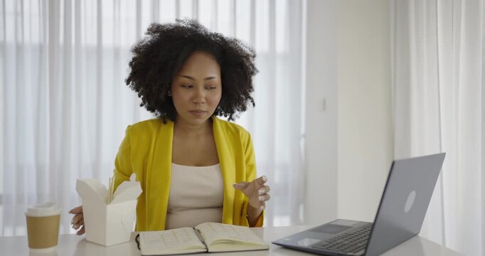 Portrait of young African woman enjoying Chinese food in office lunch break, female employee holding chopsticks eating takeaway Chinese or Korean noodles lunch box meal at workplace in home office.