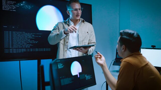 Two young male IT professionals brainstorming and analyzing AI system programming code while working with multiple computer screens and digital tablet in a high tech office