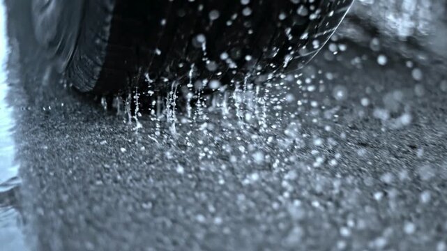 Close up view of car tire spinning rapidly on wet asphalt road during heavy rain creating water splashes on surface in slow motion for automotive background.