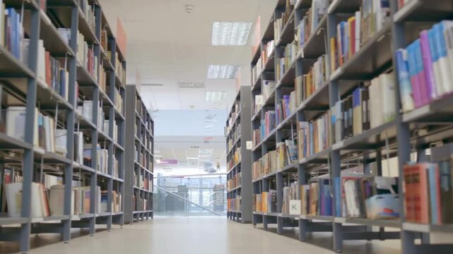 Smooth tracking shot through a modern library aisle with tall bookshelves packed with colorful books, revealing a bright, contemporary reading area beyond.