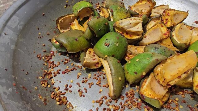 Spiced green mango slices with chili flakes, traditional raw mango pickle preparation in metal bowl, sour fruit snack with red pepper seasoning