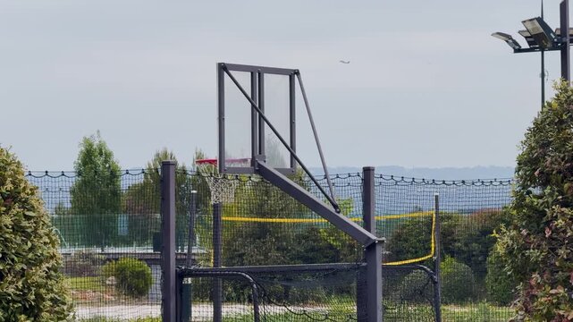 Outdoor basketball hoop in a fenced garden court near the sea, showing a private sports area with netting, trees and coastal scenery.