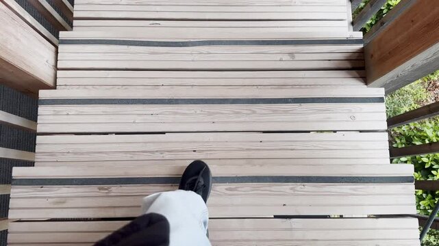 Point-of-view shot of a person walking down wooden outdoor stairs, showing deck boards, black shoes and railing in a rustic exterior setting.