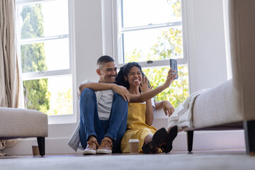 Fototapeta premium Couple sitting on floor by large windows taking selfie with smartphone and to-go cups nearby