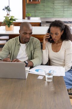 Diverse couple sitting at dining table in kitchen, using laptop and smartphone, reviewing charts