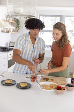 Diverse couple standing at white kitchen island preparing and plating pancakes with spatula