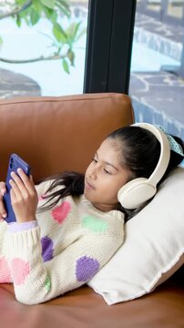 Vertical video: Shifting child in sweater on tan sofa reaching blue phone donning headset watching
