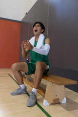 Naklejka premium Basketball player sitting on bench, clapping and shouting at court, towel and blue bottle nearby