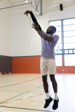 Male basketball player is jumping on wood court in gym, following through toward hoop and backboard
