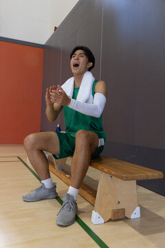 Basketball player sitting on bench, clapping and shouting at court, towel and blue bottle nearby