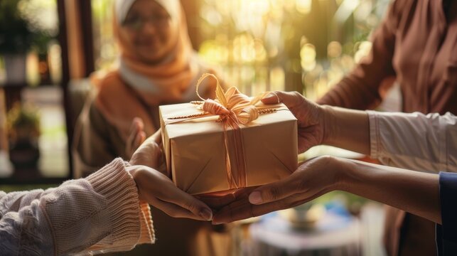 Hands exchanging a beautifully wrapped gift box with a ribbon, with a blurred background of people celebrating in a warm, inviting indoor setting