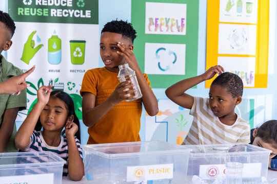 Diverse school-age children sorting recycling at classroom table, labeled PLASTIC bins and bottle