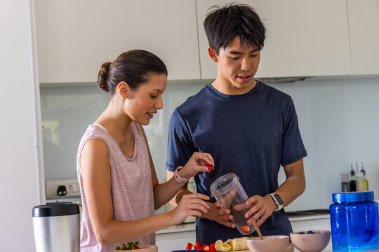 Diverse couple placing strawberries into clear blender cup at home kitchen counter, holding knife