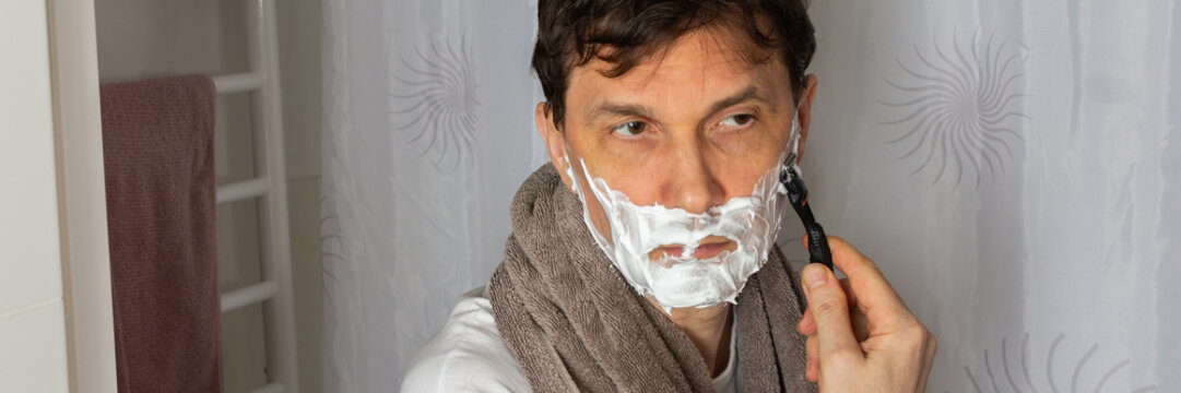 A 53 year old man shaves in the bathroom during his morning routine. He uses a razor and shaving cream while wearing a towel around his neck. Water is visible in the sink, banner