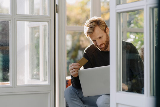 Man using laptop and credit card in sunroom at home