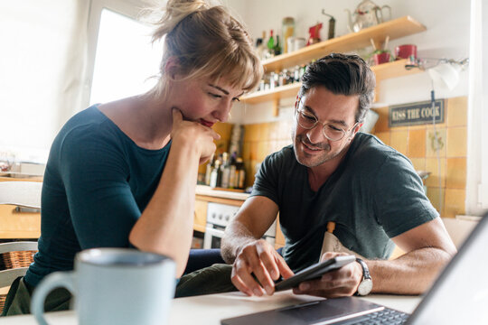 Couple sitting at table in kitchen using smartphone