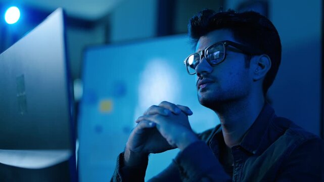 Focused young male professional with glasses working late at a computer in a dark blue room. Concentrated software developer or data analyst analyzing information on a screen during night hours.