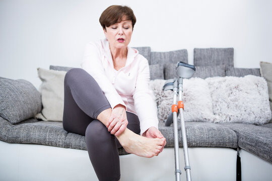 Senior woman with crutches sitting on couch, checking her ankle