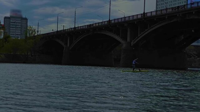 A man is sailing on a sapboard along a river under a large city bridge against a background of cloudy sky, water and a city embankment.
