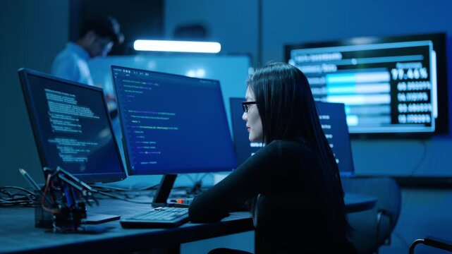Young asian female programmer writes code on multiple monitors in a dark, high-tech cybersecurity center. IT specialist analyzes software data late at night. Modern tech office with a male colleague.