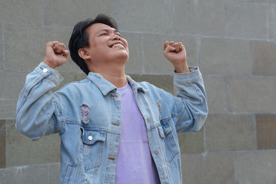 Excited Young Asian Man Celebrating Success with Raised Fists Against a Stone Wall