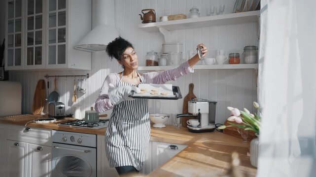 Black woman taking selfie in home kitchen, holding baking sheet with cookies. African american lady photographing herself by smartphone for memories and posting on social media, culinary blogger