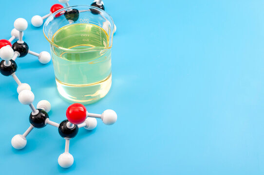 Ball-and-stick ethanol molecule model beside a glass beaker of alcohol liquid on a blue background with copy space. Organic chemistry, biofuel and science education concept with text space