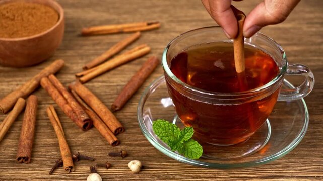 Hand stirring hot herbal tea with a cinnamon stick in a clear glass cup on a rustic wooden table
