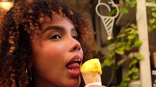 Gorgeous young black woman with curly hair enjoying a sweet moment as she sensually licks a delicious yellow ice cream cone outside a gelateria during a warm summer evening