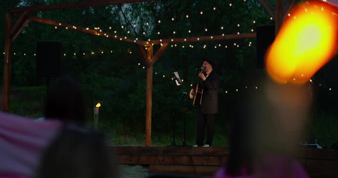 Male Musician Singing with Acoustic Guitar on Small Outdoor Stage at Dusk