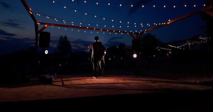 Silhouette of man on outdoor wooden stage under string lights at dusk