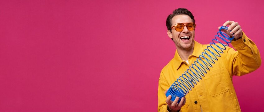 Happy man playing slinky toy wearing yellow jacket and orange sunglasses on pink background, laughing millennial male posing with retro plastic spring coil, cheerful vibrant studio portrait with space