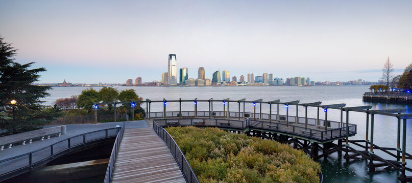 Wooden boardwalk promenade leads to a panoramic waterfront view of the Jersey City skyline from a park in Lower Manhattan, highlighting a popular urban tourism destination, New York City.